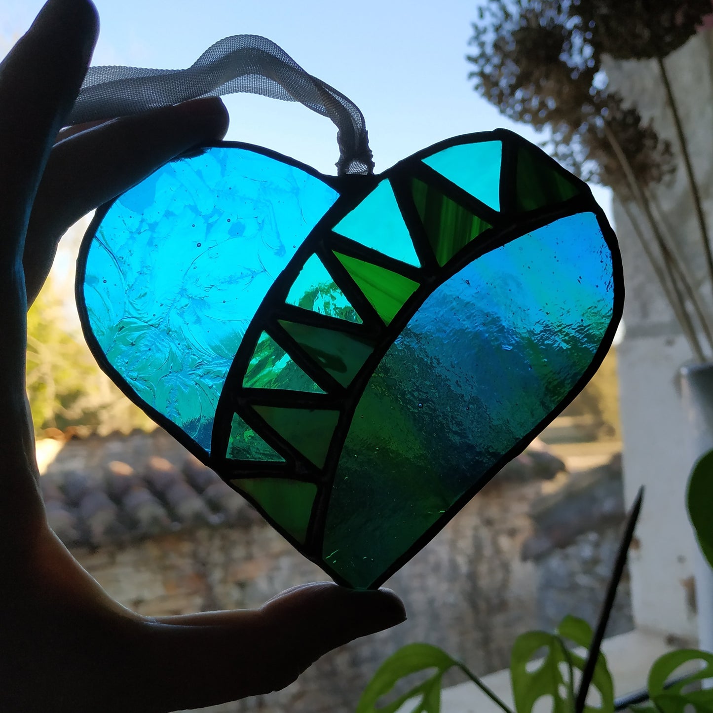 Silhouette of hand holding a satined glass heart made by Pamela Angus up to a window 